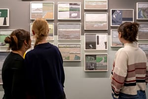 Drei junge Frauen betrachten Gemälde in einer Kunstausstellung, die aus mehreren kleinformatigen Bildern besteht, ordentlich an der Wand arrangiert.